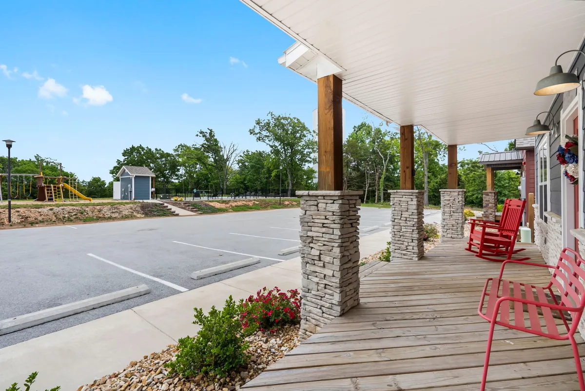 A cozy exterior view showing red rocking chairs lined up on a porch, overlooking a playground in the background. The scene invites relaxation for adults while kids enjoy safe play nearby; the stone pillars and landscaping add a touch of elegance and warmth, echoing the boutique resort vibe.