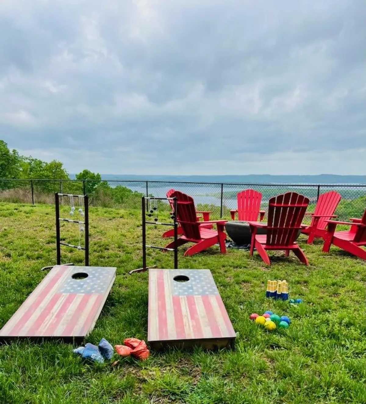 Focusing on red Adirondack chairs encircling a fire pit, with cornhole boards set up nearby and a view of the water in the background, this image highlights spaces designed for family games, relaxation, and evening gatherings under the open sky.
