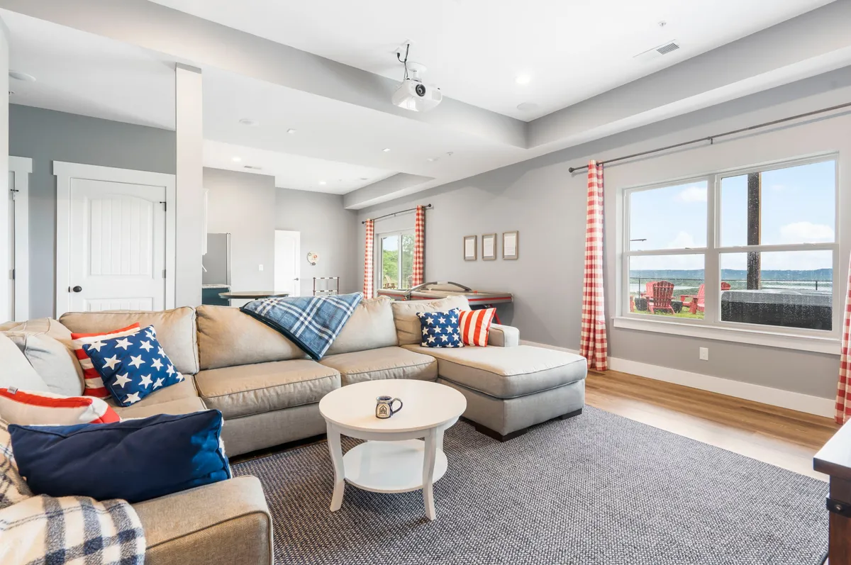 A spacious movie and entertainment room featuring a plush sectional sofa, large windows with scenic views, and a ceiling-mounted projector. Decorative pillows and relaxed lighting create a cozy atmosphere ideal for group lounging and movie nights.