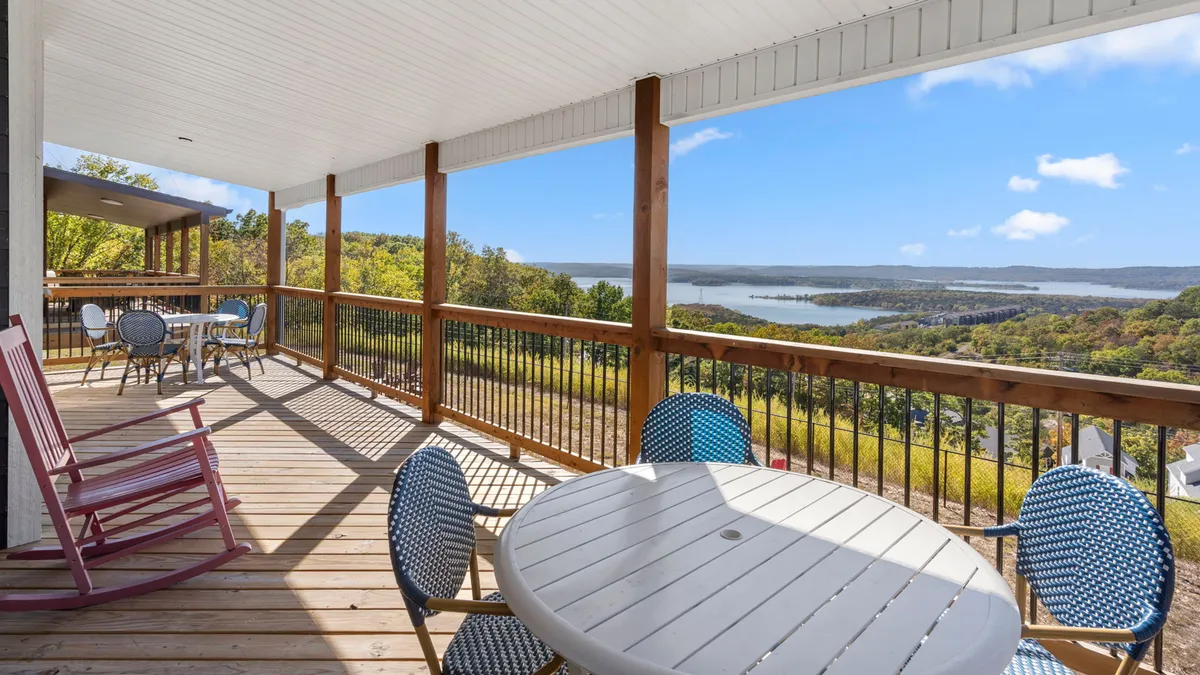 A wooden deck with colorful chairs overlooks the serene lake, creating a seamless transition from the interior gathering spaces to the outdoors. The inviting setup suggests easy access for relaxing, entertaining, or soaking in the stunning lake views.