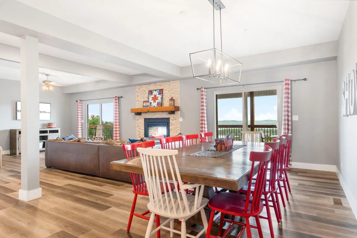 A spacious, open-concept dining and living area featuring a large dining table, a double-sided fireplace, and generous windows offering glimpses of the outdoors. The setup encourages both cozy meals and relaxed gatherings by the fire, with a warm, welcoming ambiance throughout.