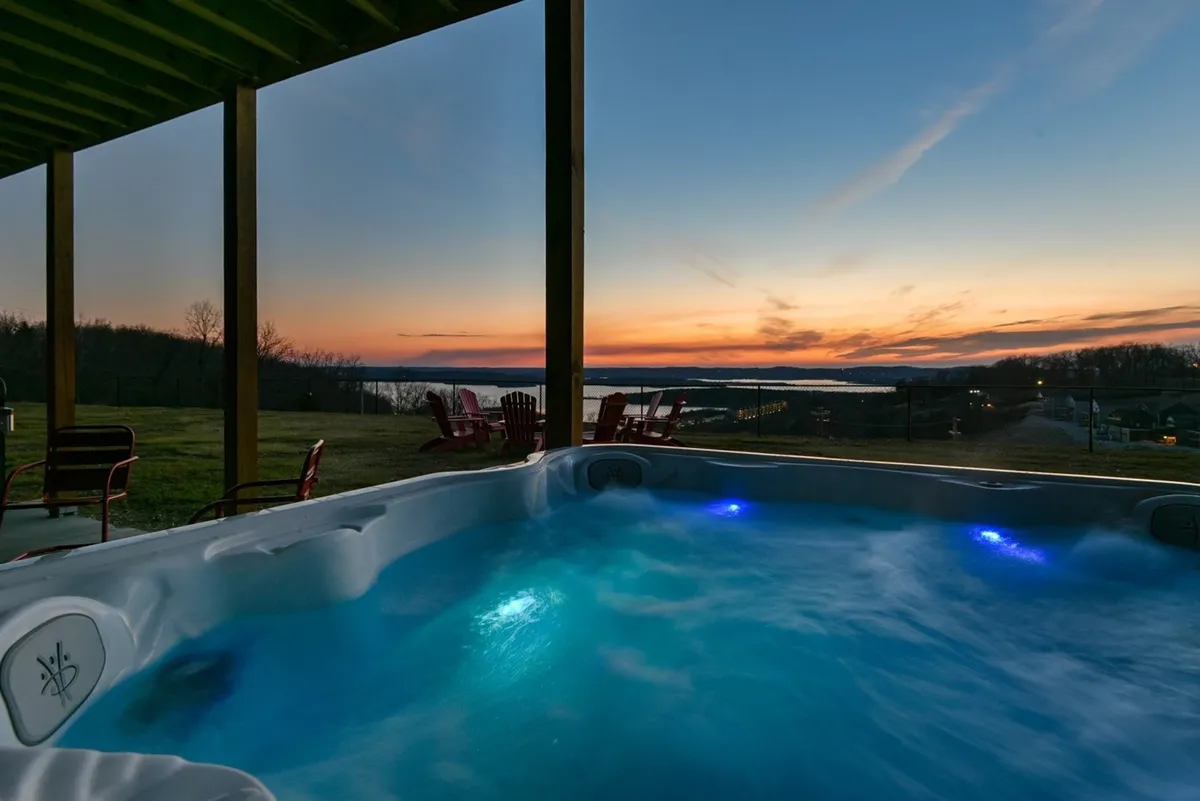 An inviting exterior featuring a hot tub surrounded by Adirondack chairs with a tranquil water view and a glowing sunset. The setup encourages socializing, stargazing, and soaking outdoors in a private, scenic setting.
