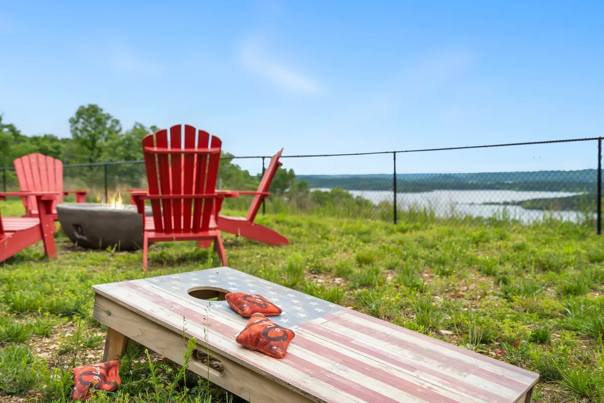 A fun lakeside setting with red Adirondack chairs encircling a fire pit, a cornhole game set up nearby, and a scenic view of the lake. The atmosphere is casual and perfect for socializing into the evening.