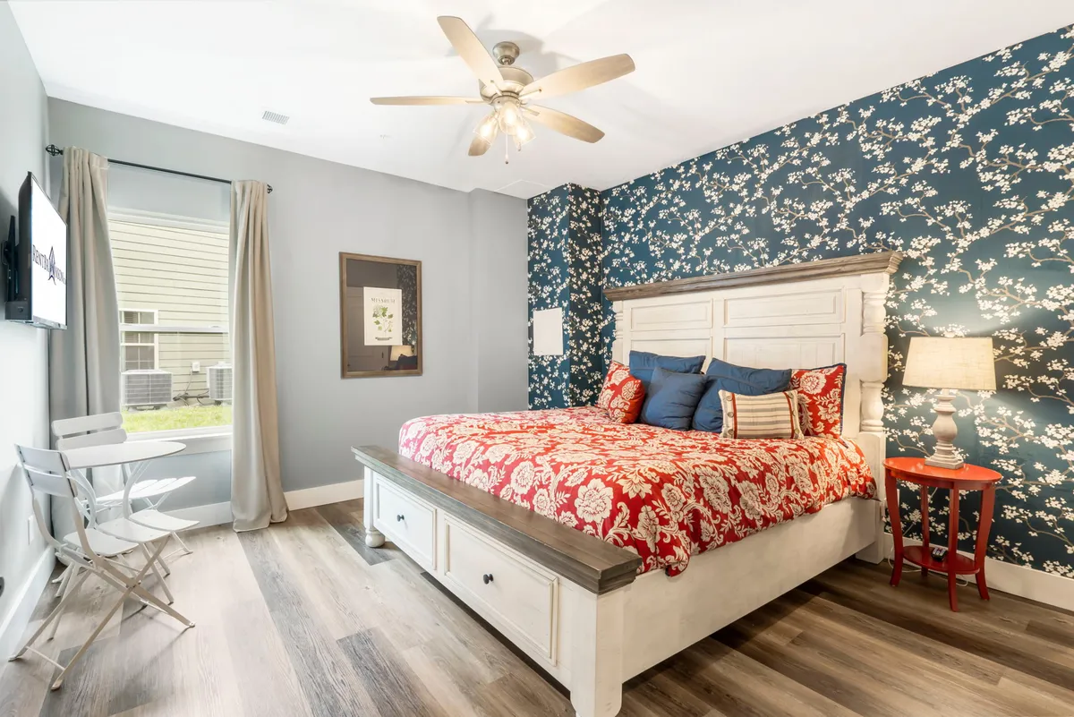 A bright, comfortable bedroom featuring a plush bed with soft linens, decorative wallpaper, a side table with a lamp, and a window allowing natural light to pour in. The atmosphere is calm, inviting, and well-suited for relaxation after a long day.